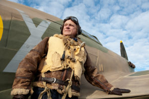 Low angle view of a World War Two RAF pilot with his Hurricane fighter aircraft against a blue sky with clouds. The pilot is wearing his famous Irvin leather flying jacket and "Mae West" life jacket.