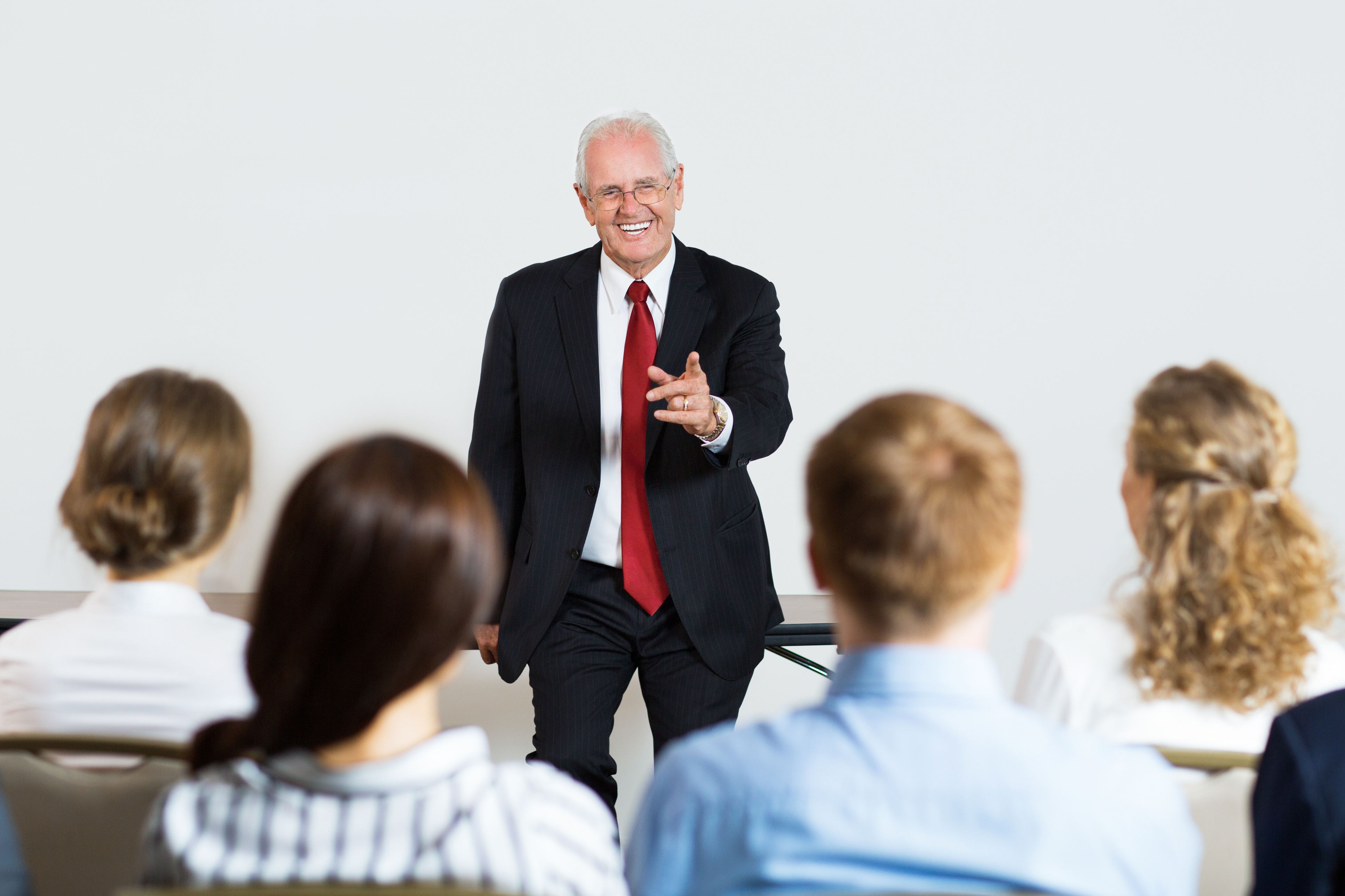Smiling senior businessman leaning on table and pointing to sitting audience of four people. Rear view of audience.